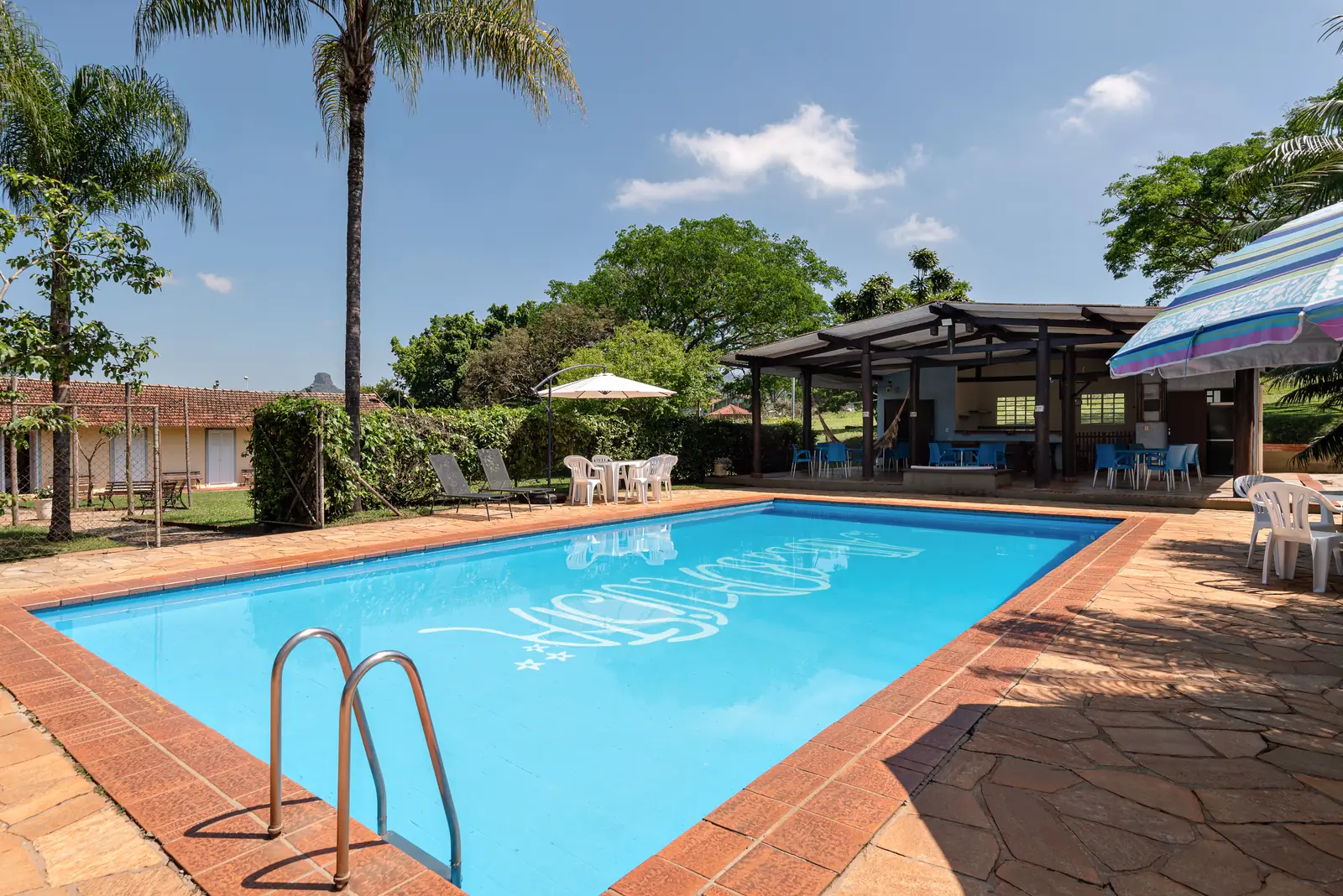 Piscina com o logo do hotel e o quiosque ao fundo, sob céu azul